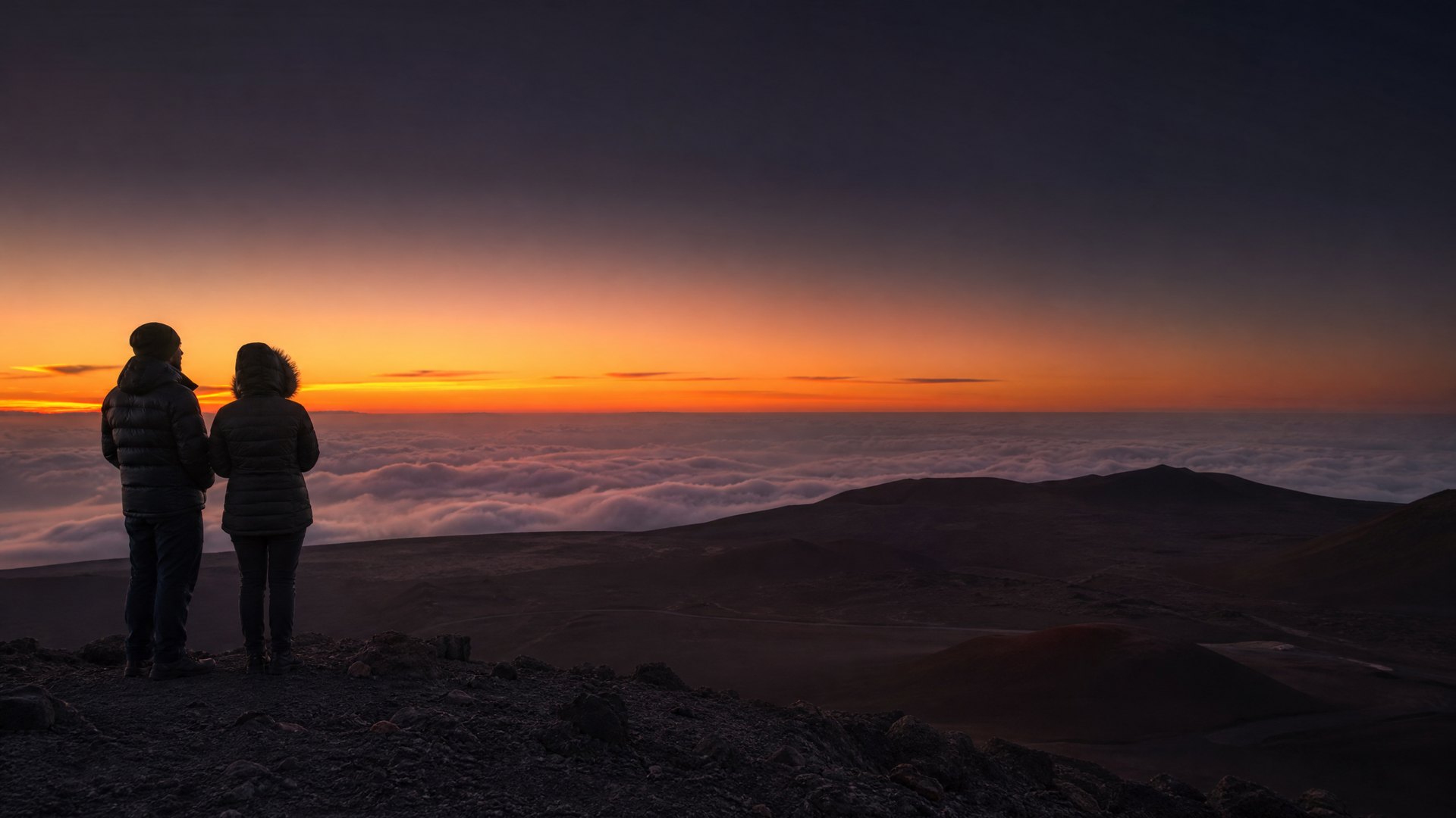 Two visitors in parkas watching the sunset above clouds on Mauna Kea